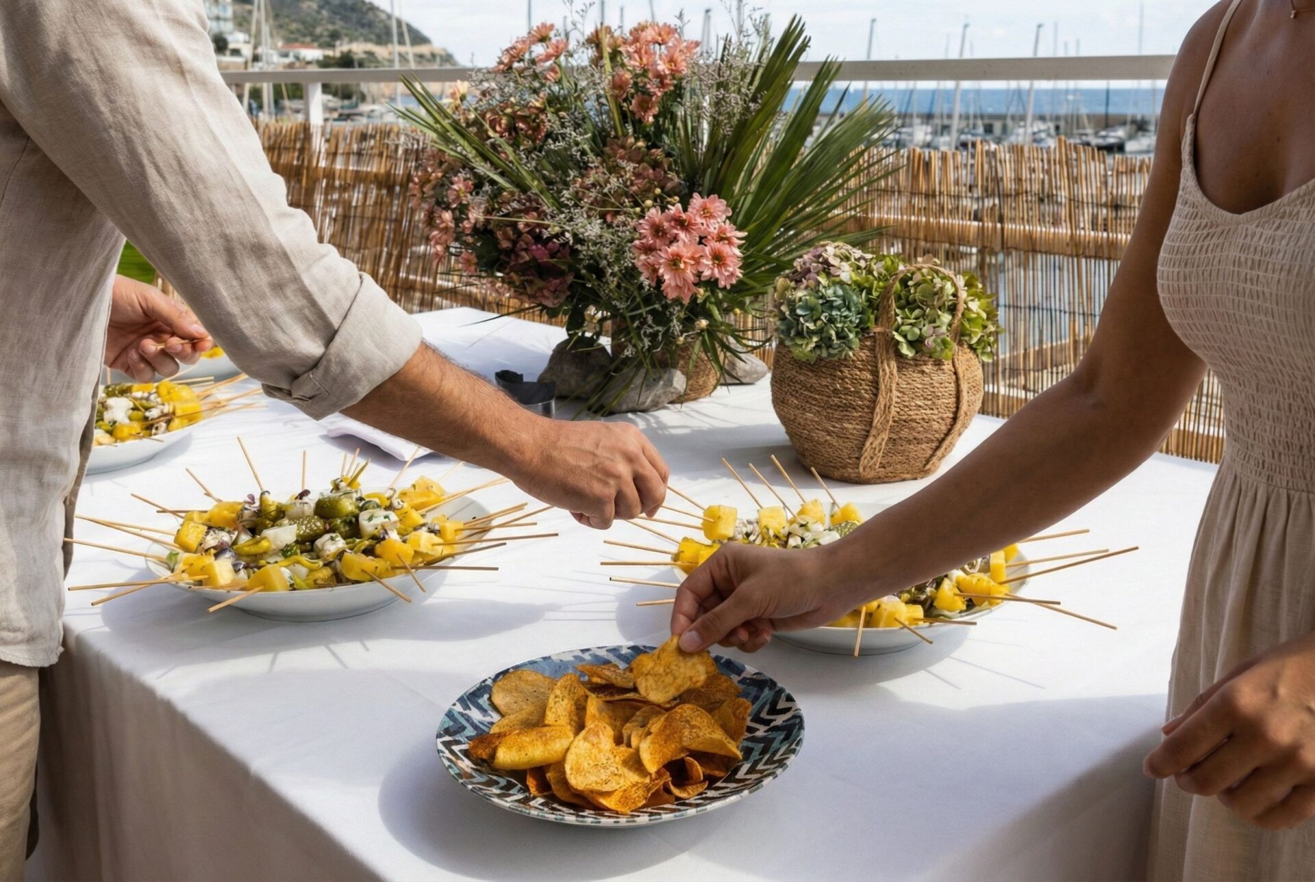 Detalle de invitados cogiendo brochetas y aperitivos frescos en una mesa decorada frente al puerto deportivo de El Nàutic.