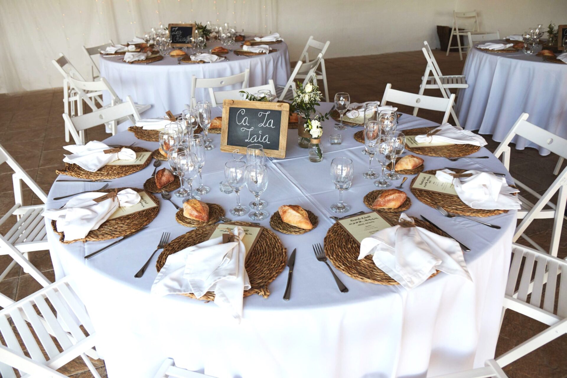 Detalle de mesa para boda rústica con bajoplatos de mimbre, sillas blancas plegables y pizarra decorativa en masía restaurante cerca de Barcelona.